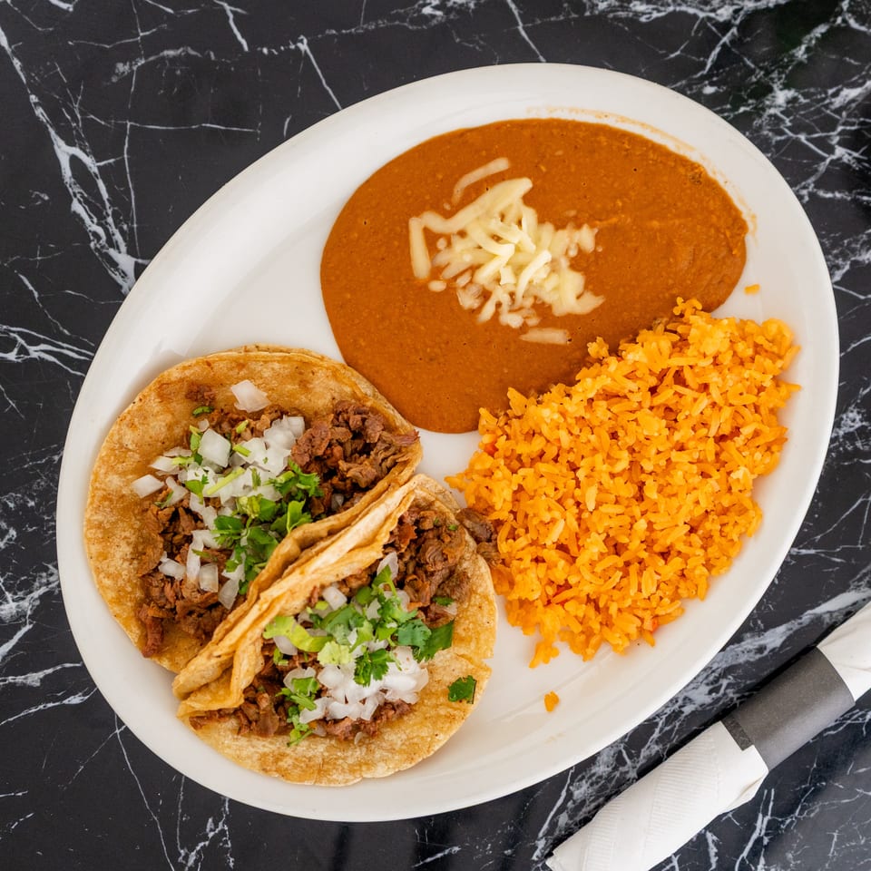 Two tacos with rice and refried beans on a white plate, on a black marble table.