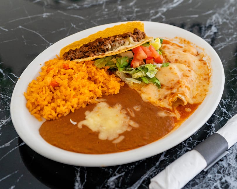A white plate with a hard-shell taco, rice, refried beans, and two cheese enchiladas.