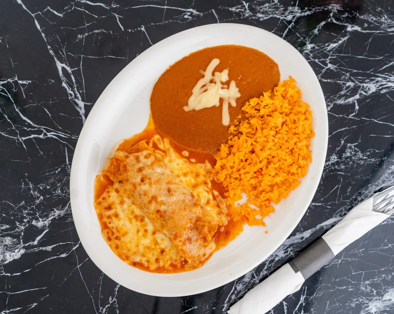 A plate of enchiladas with melted cheese, refried beans, and orange rice on a black marble table.