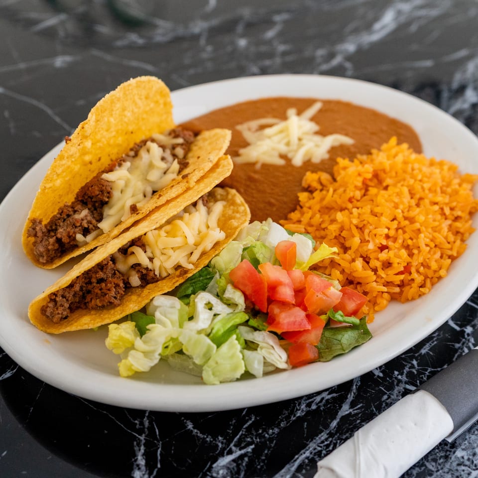 Two beef tacos with cheese, refried beans, rice, and a side salad on a white plate.