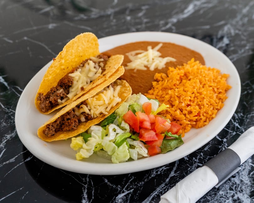 Two beef tacos with cheese, refried beans, rice, and a side salad on a white plate.