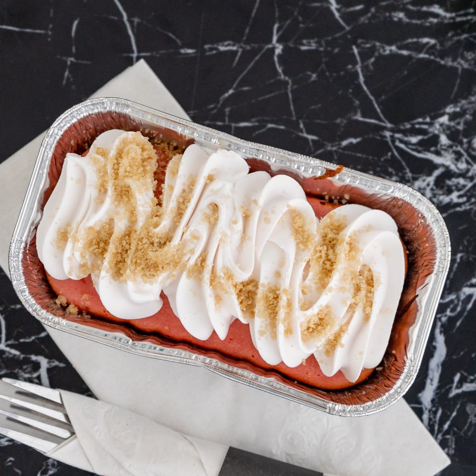 Red velvet cake in a foil pan, topped with white cream and brown sprinkles, on a marble surface.