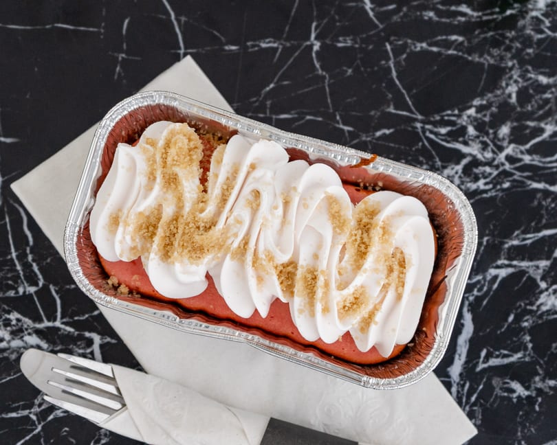 Red velvet cake in a foil pan, topped with white cream and brown sprinkles, on a marble surface.