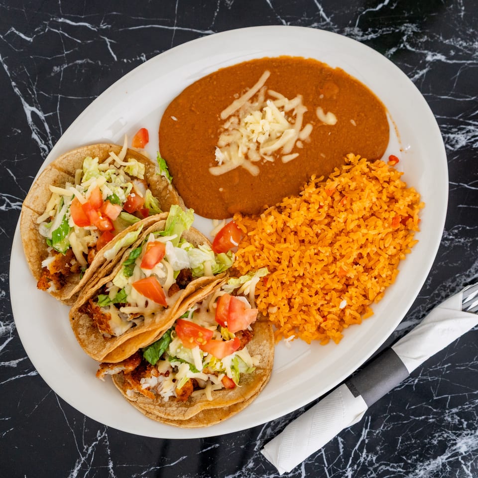 Three tacos with lettuce, tomato, and cheese, alongside refried beans and orange rice on a white plate.