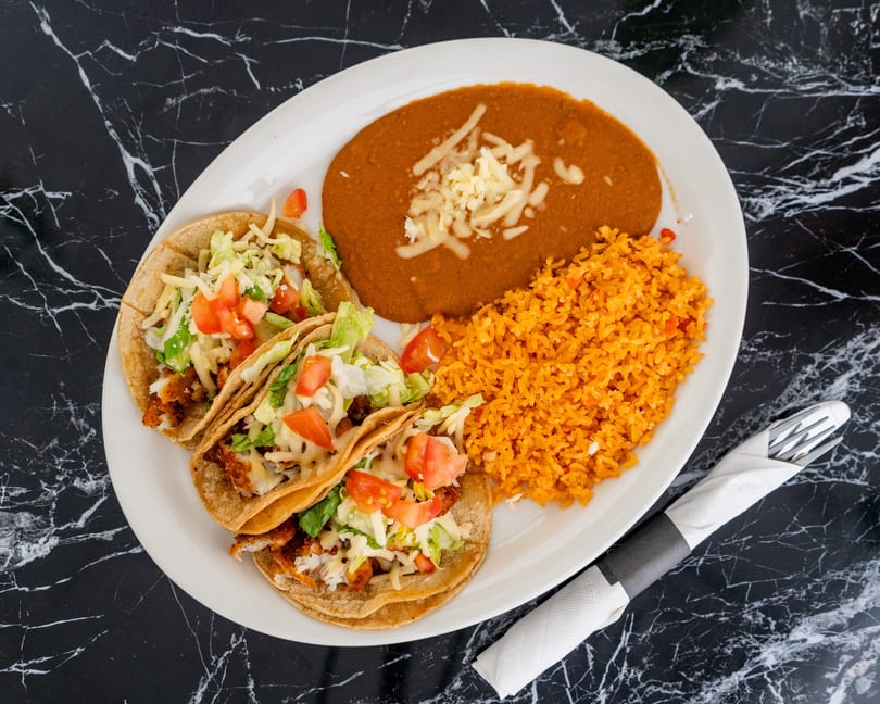 Three tacos with lettuce, tomato, and cheese, alongside refried beans and orange rice on a white plate.