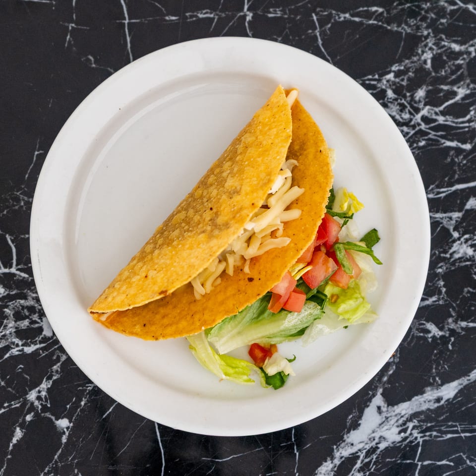 A single hard-shell taco filled with meat, shredded cheese, lettuce, and diced tomatoes on a white plate.
