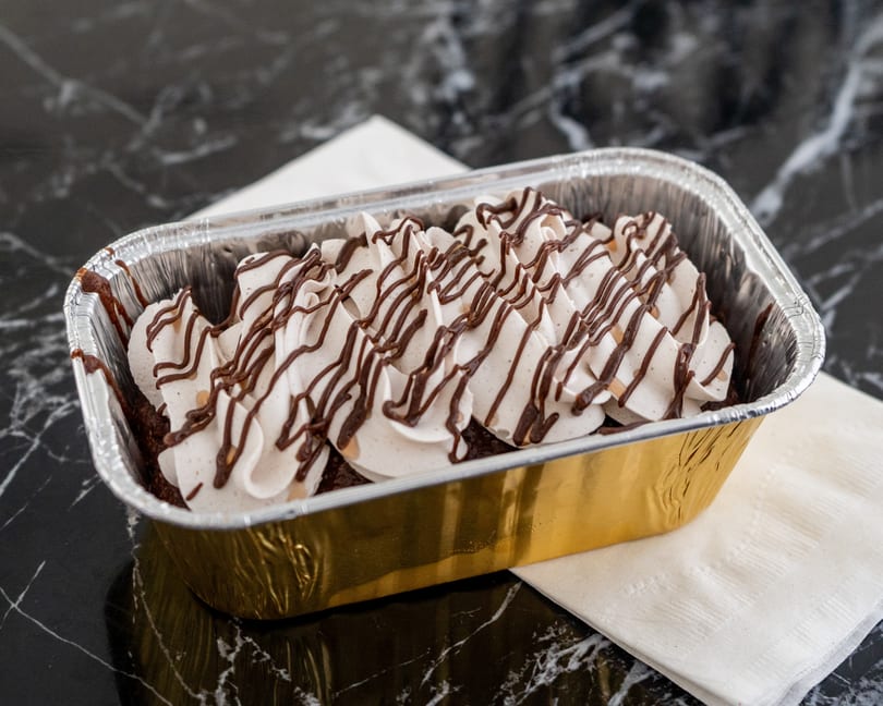 Chocolate cake with white whipped cream and chocolate drizzle in a gold and silver foil pan.